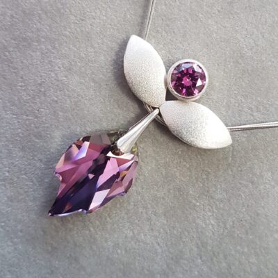 Silberner Engel mit funkelndem Violett Silberner Engel-Anhänger-Schmuck mit zwei flügelartigen Elementen und einem runden, violetten Stein oben, daran hängend ein facettierter, tropfenförmiger Kristall in Violett- und Lilatönen auf grauem Hintergrund