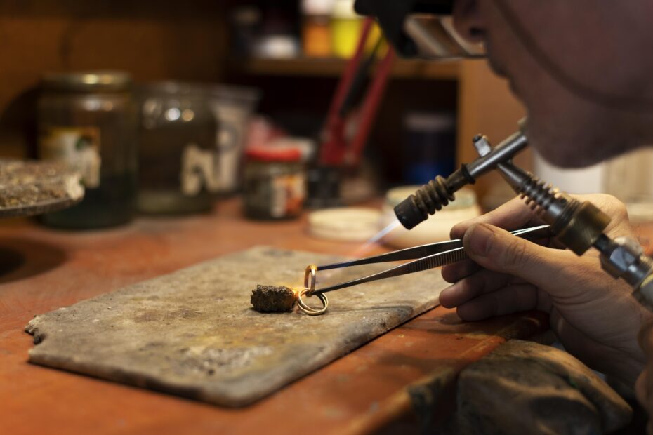 Ein Goldschmied arbeitet an einem Ring mit einer Lötlampe und Zangen auf einer Werkbank aus Holz.