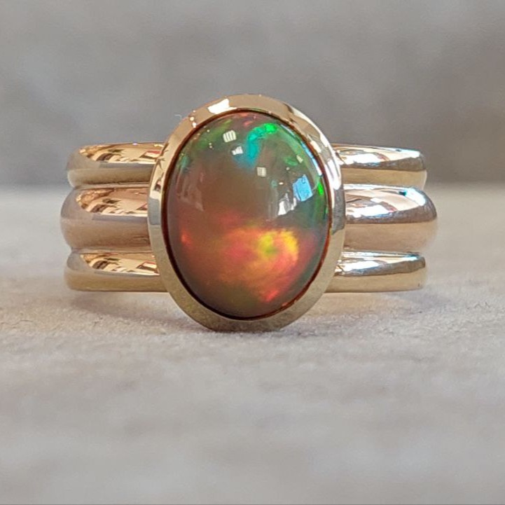 Goldring mit einem ovalen Opal, der grüne und rote Farbreflexe zeigt, auf einem hellen Untergrund.